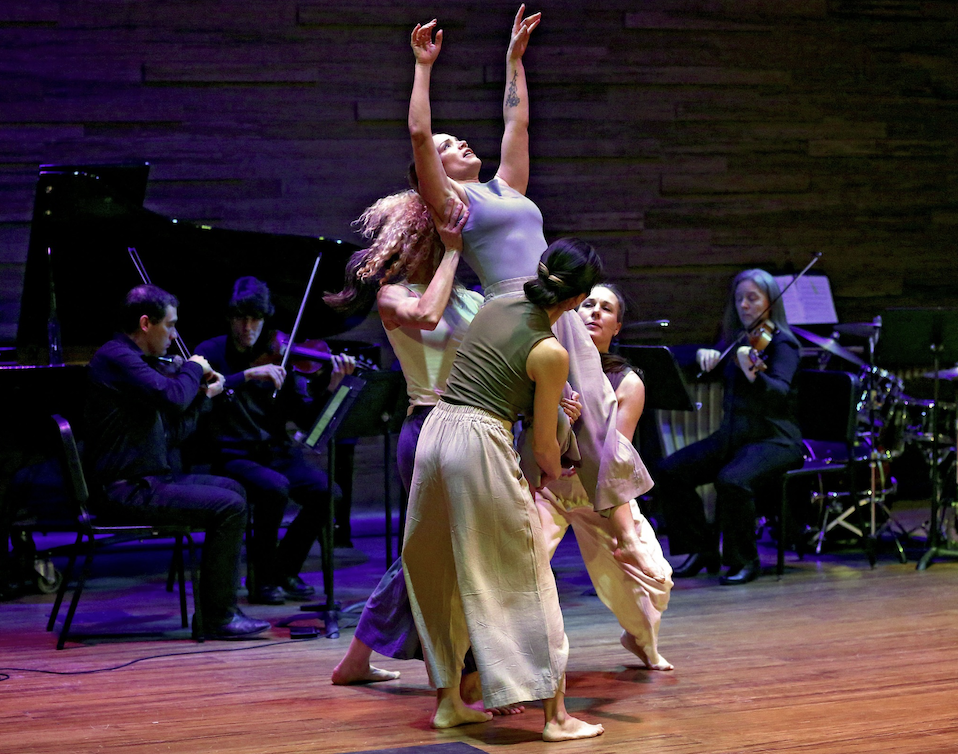 Photo of two female dancers lifting a third female dancer, with musicians playing in the background, by Royce Burgess